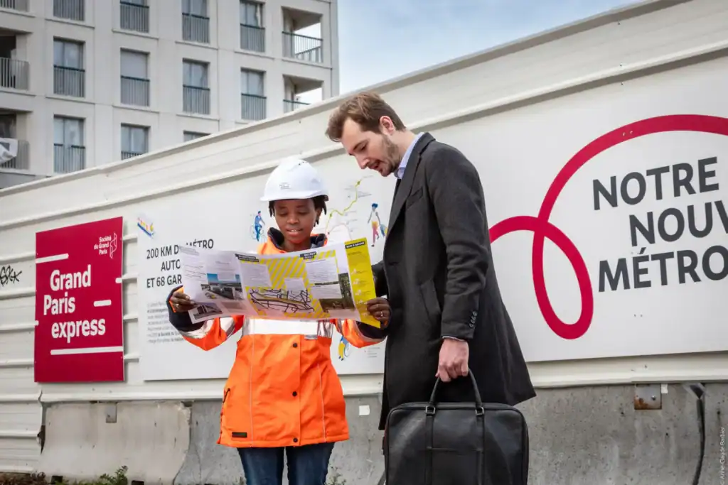Deux professionnels discutent devant un panneau du Grand Paris Express sur un chantier.